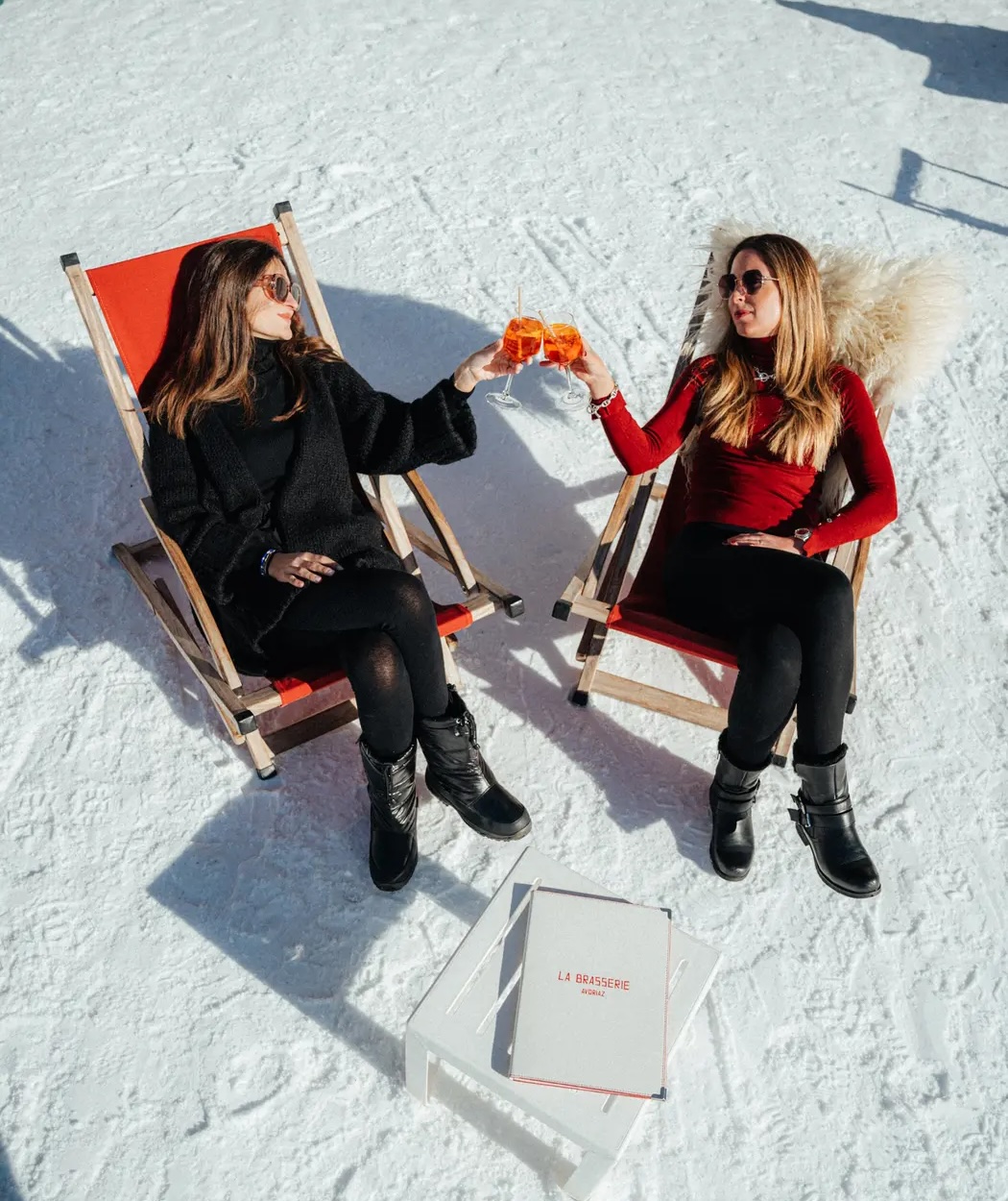 Deux femmes portant des lunettes de soleil, assises sur des chaises longues rouges dans la neige, trinquent avec des boissons orange, restaurant La Brasserie à Avoriaz, Haute-Savoie, France.