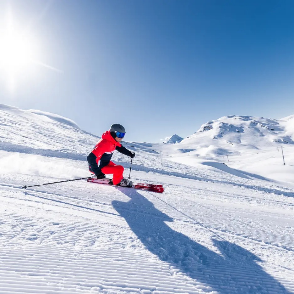 Skieur en tenue rouge effectuant un virage coupé sur une piste enneigée, avec des montagnes et un ciel bleu limpide en arrière-plan, restaurant La Brasserie à Avoriaz, Haute-Savoie, France.