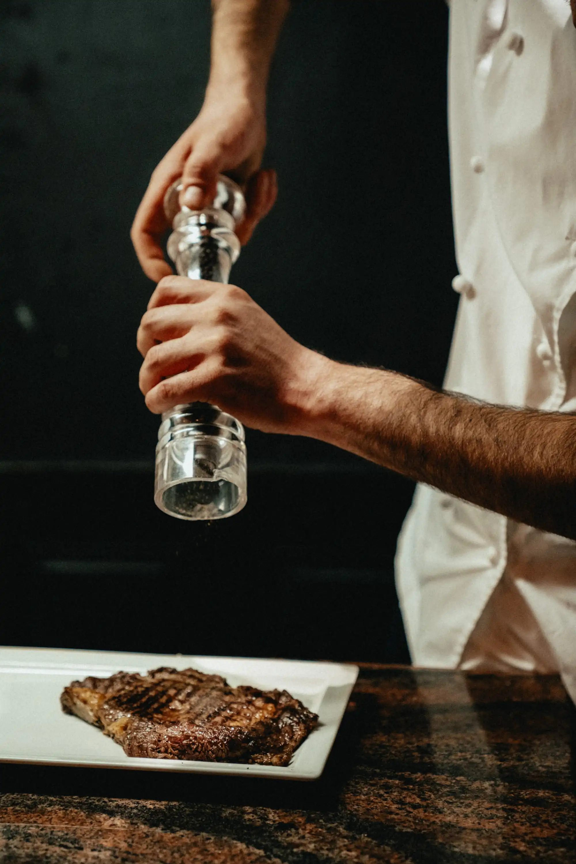 Un chef assaisonne un steak grillé dans une assiette blanche à l'aide d'un moulin à poivre, au-dessus d'un plan de travail sombre, restaurant La Brasserie à Avoriaz, Haute-Savoie, France.