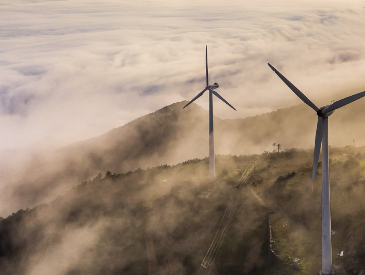 Wind turbines on a mountain partially covered by fog with clouds in the background at sunrise or sunset.