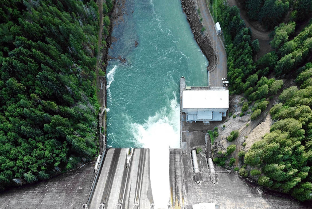 Aerial view of a dam releasing water with forested land on both sides of the river.