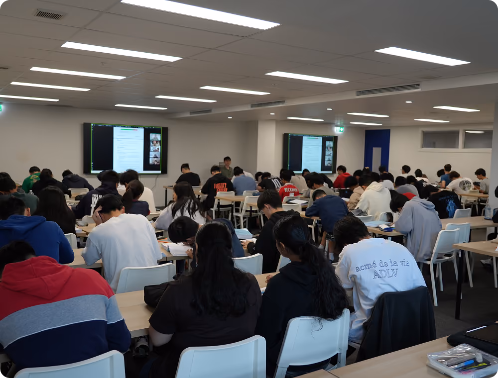 A wide classroom photograph showing many students seated at desks and facing a tutor and presentation screen during an EdAtlas lesson.
