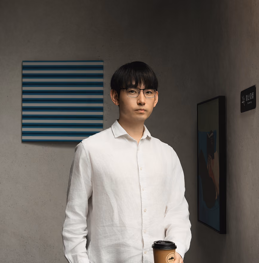 A young male EdAtlas tutor wearing a white button-up shirt stands in a classroom near a dark wall and window blinds, facing the camera.
