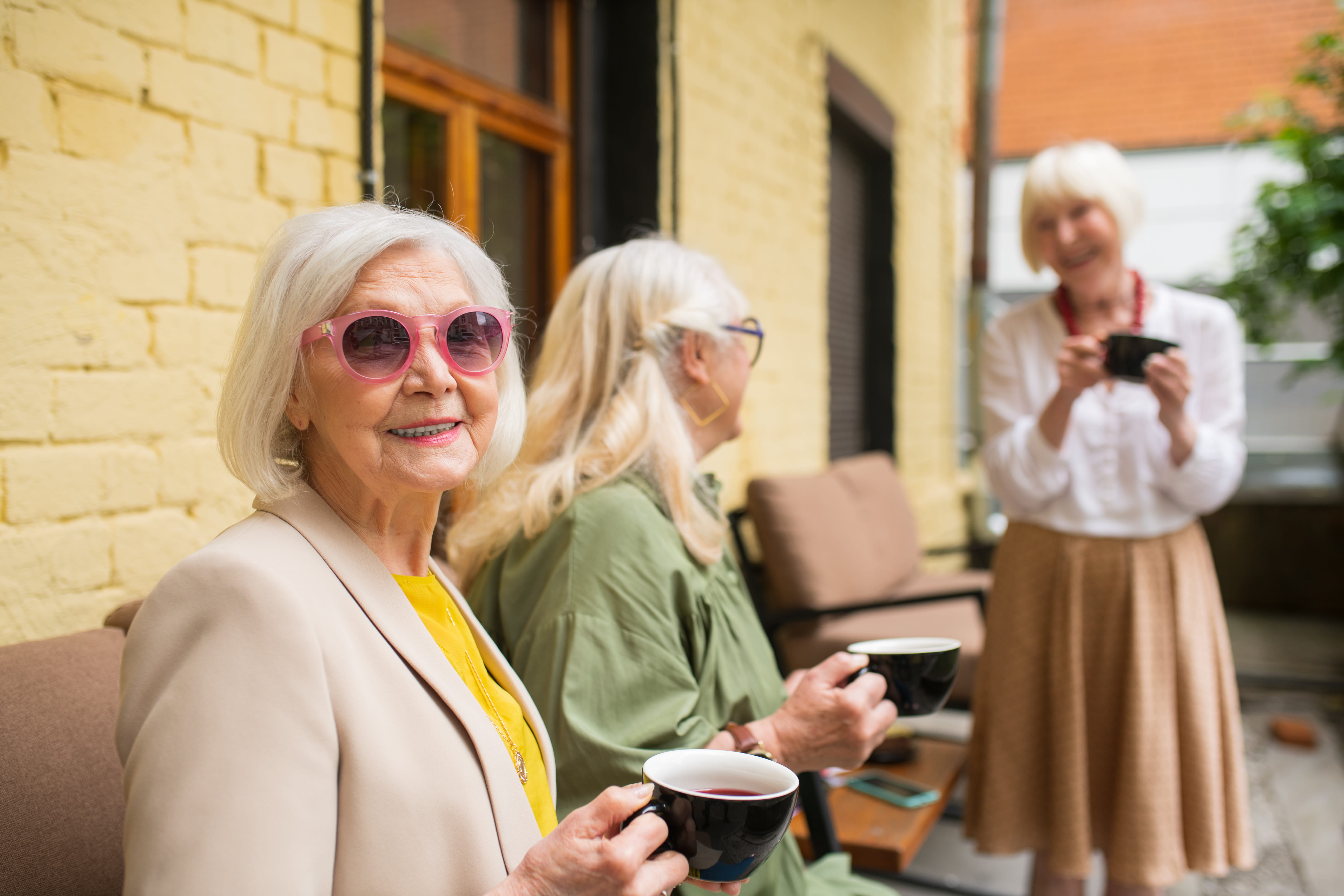 Three elderly women enjoying tea together outdoors, two sitting on a bench and one standing, all holding black cups and smiling.