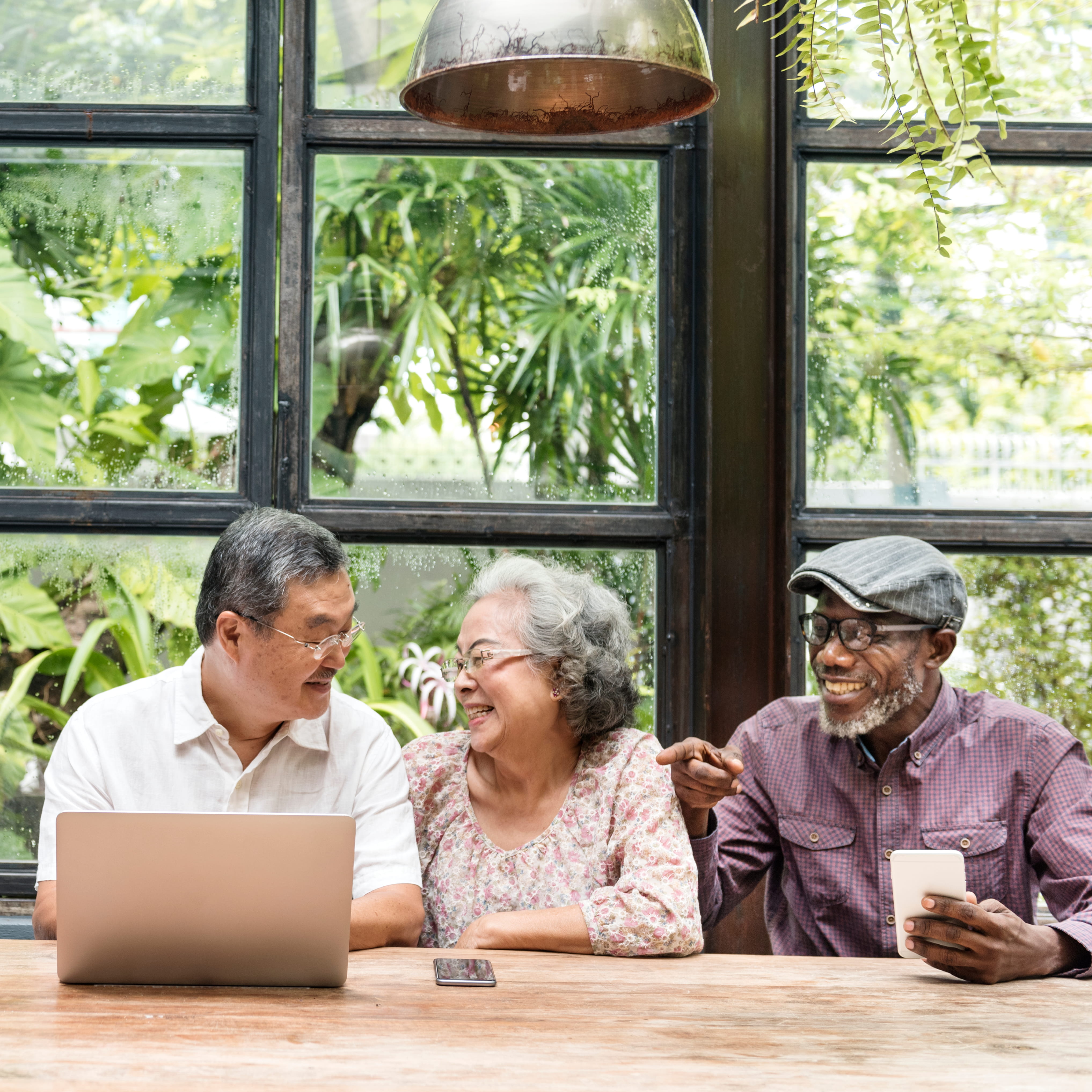 Three seniors, two men and one woman, sitting at a wooden table with a laptop and smartphone, smiling and engaging in conversation inside a bright room with large windows showing greenery outside.