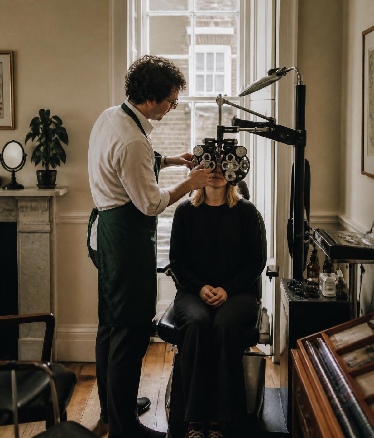 An optometrist adjusting a phoropter machine while a woman sits in a chair undergoing an eye exam in a room with a large window and wooden floor.