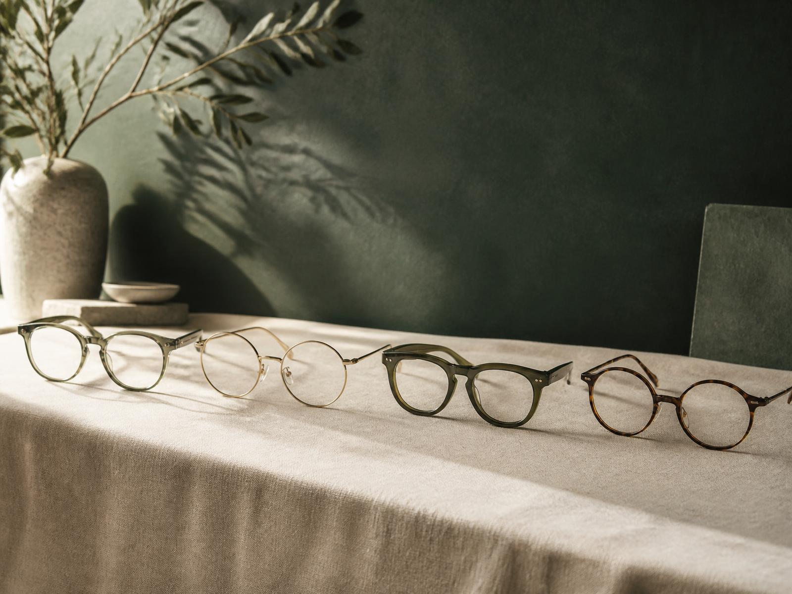 Four pairs of eyeglasses arranged in a row on a textured beige tablecloth with a green wall and a vase with leaves casting shadows in the background.