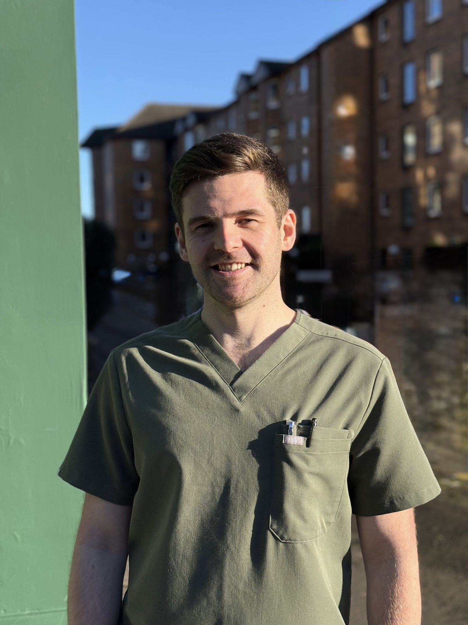 A young man in olive green scrubs standing outdoors with a slight smile, with blurred buildings and a clear blue sky in the background.