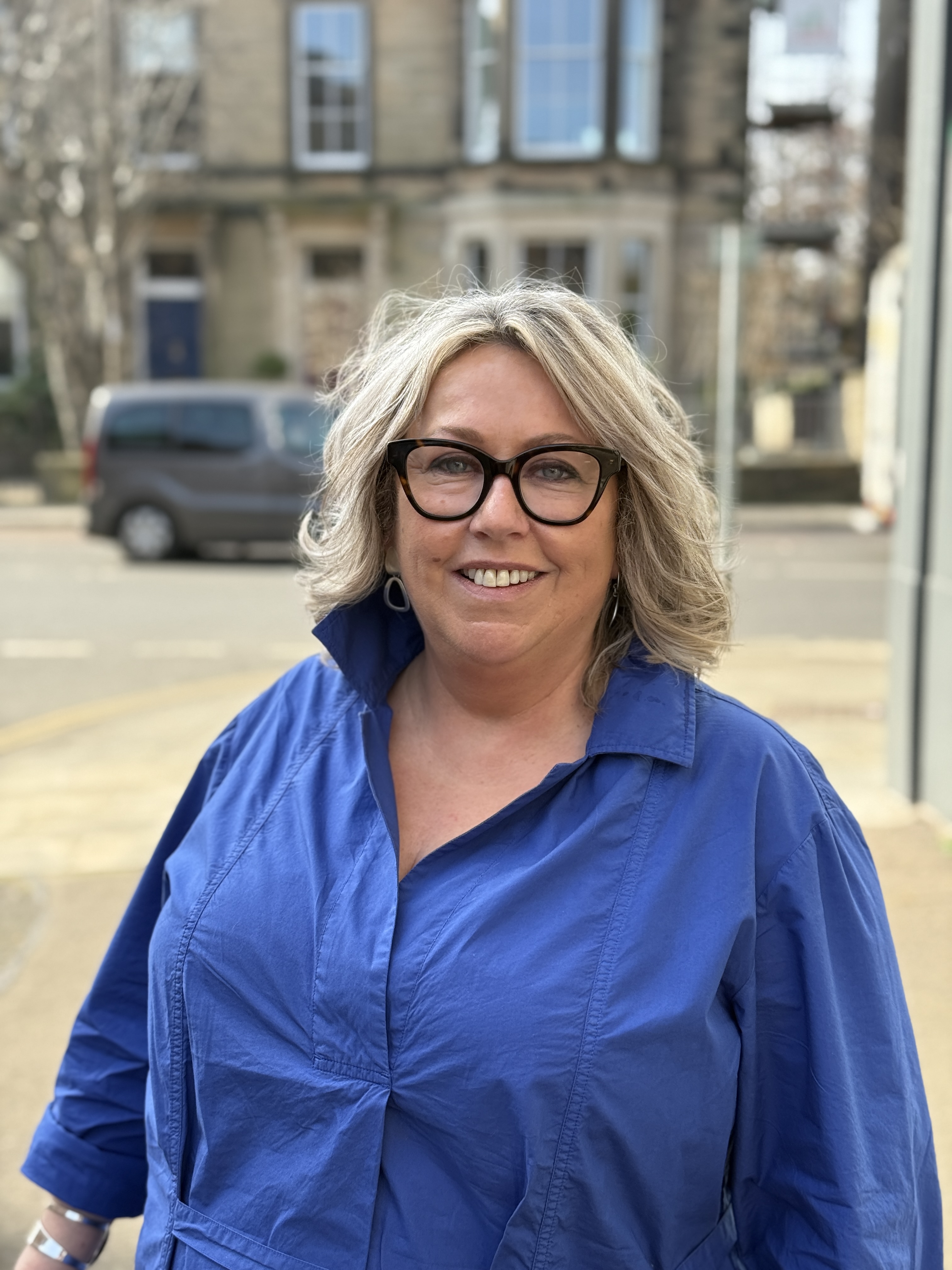 Smiling woman with blonde hair and black glasses wearing a blue collared shirt outdoors in an urban area with blurred buildings and a parked car in the background.