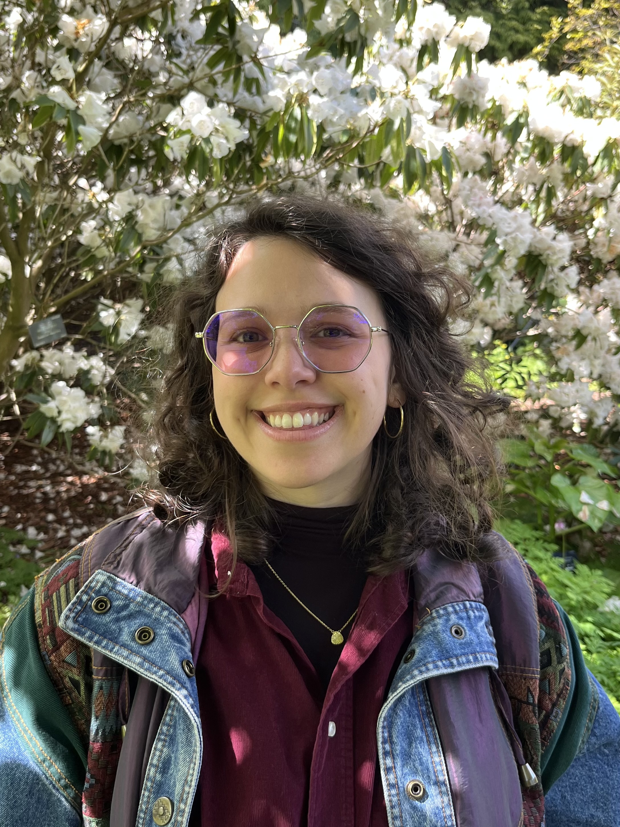 Smiling woman with curly hair wearing purple-tinted glasses, gold hoop earrings, a gold necklace, a burgundy shirt, and a layered colorful jacket, standing outdoors in front of blooming white flowers and green foliage.
