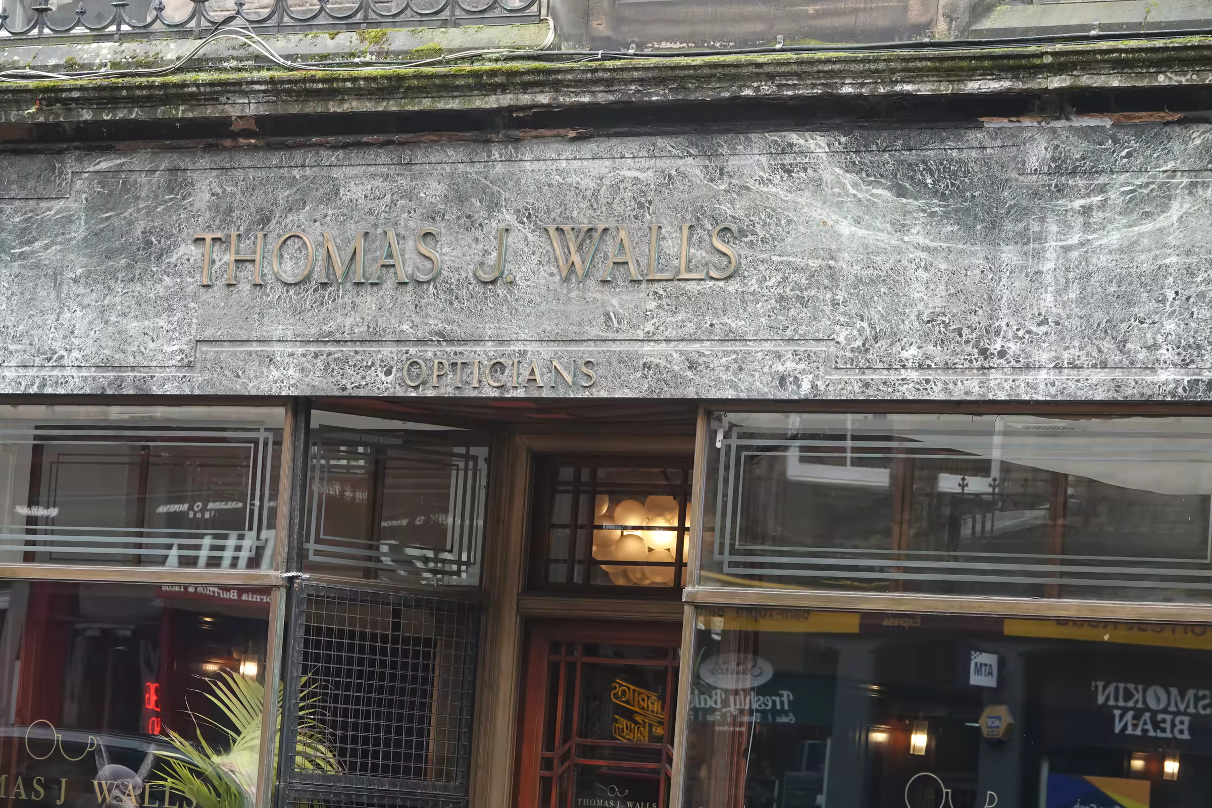 Front exterior of a building with a sign reading 'THOMAS J WALLS' and 'OPTICIANS' beneath it, featuring large glass windows and a central entrance door.