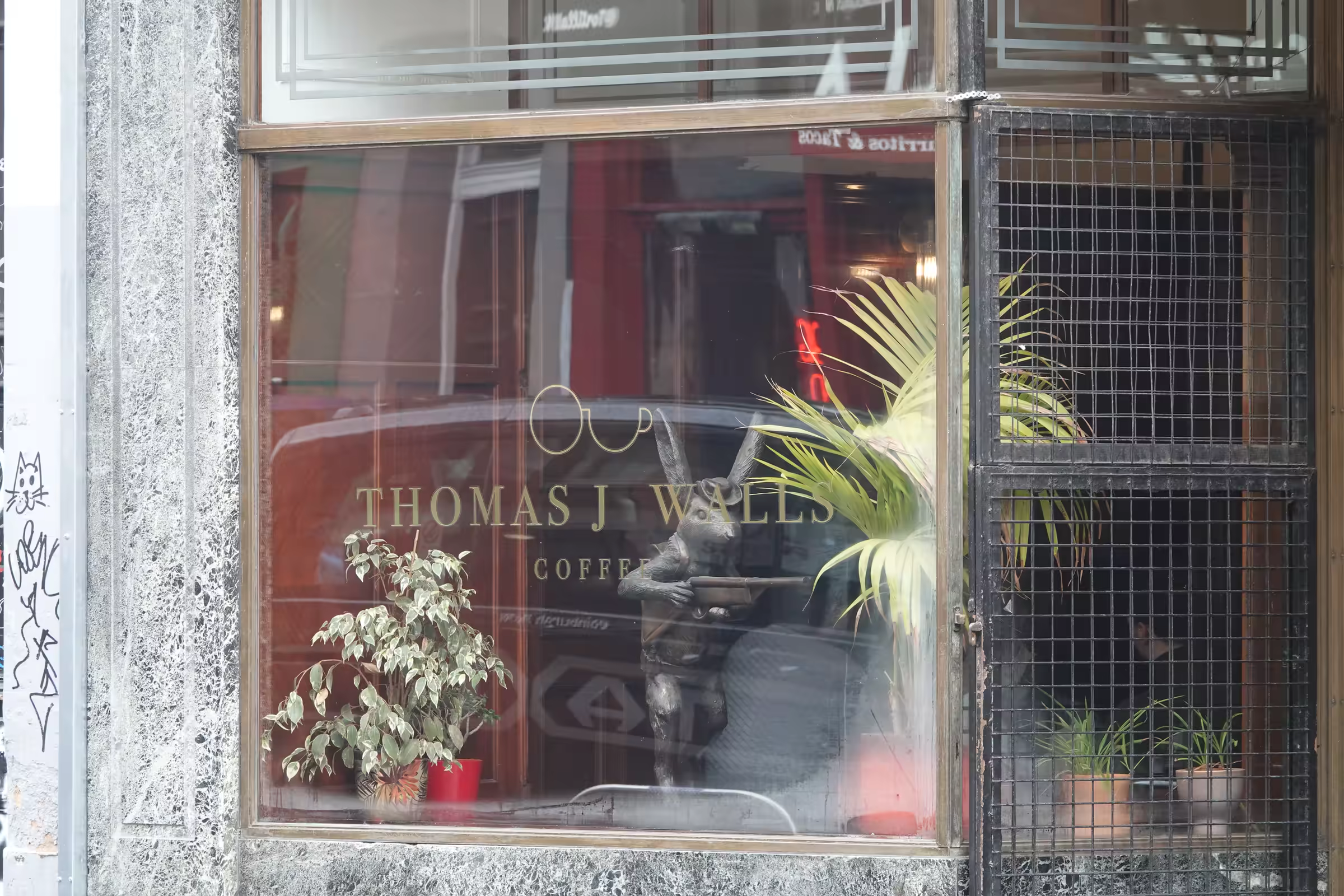 Coffee shop window with gold lettering reading 'Thomas J Walls Coffee' featuring a bronze statue of a rabbit holding a tray amidst green plants visible inside.
