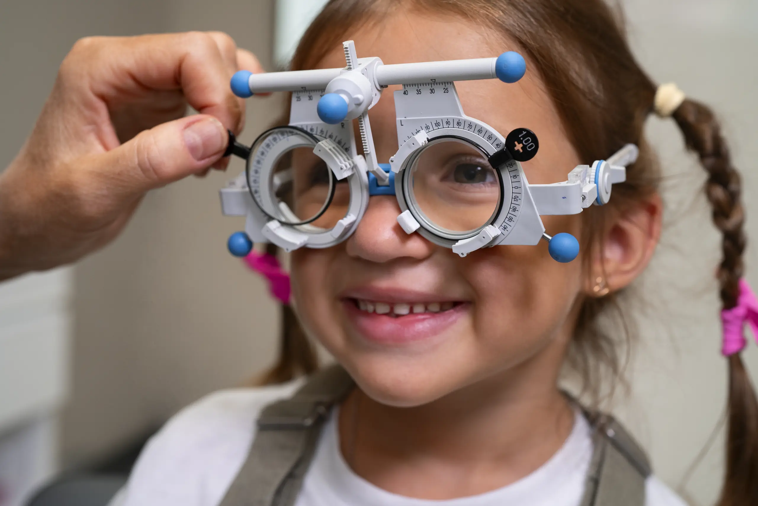 A young girl with brown hair in pigtails is smiling while an optometrist adjusts a white and blue trial frame on her face during an eye exam.