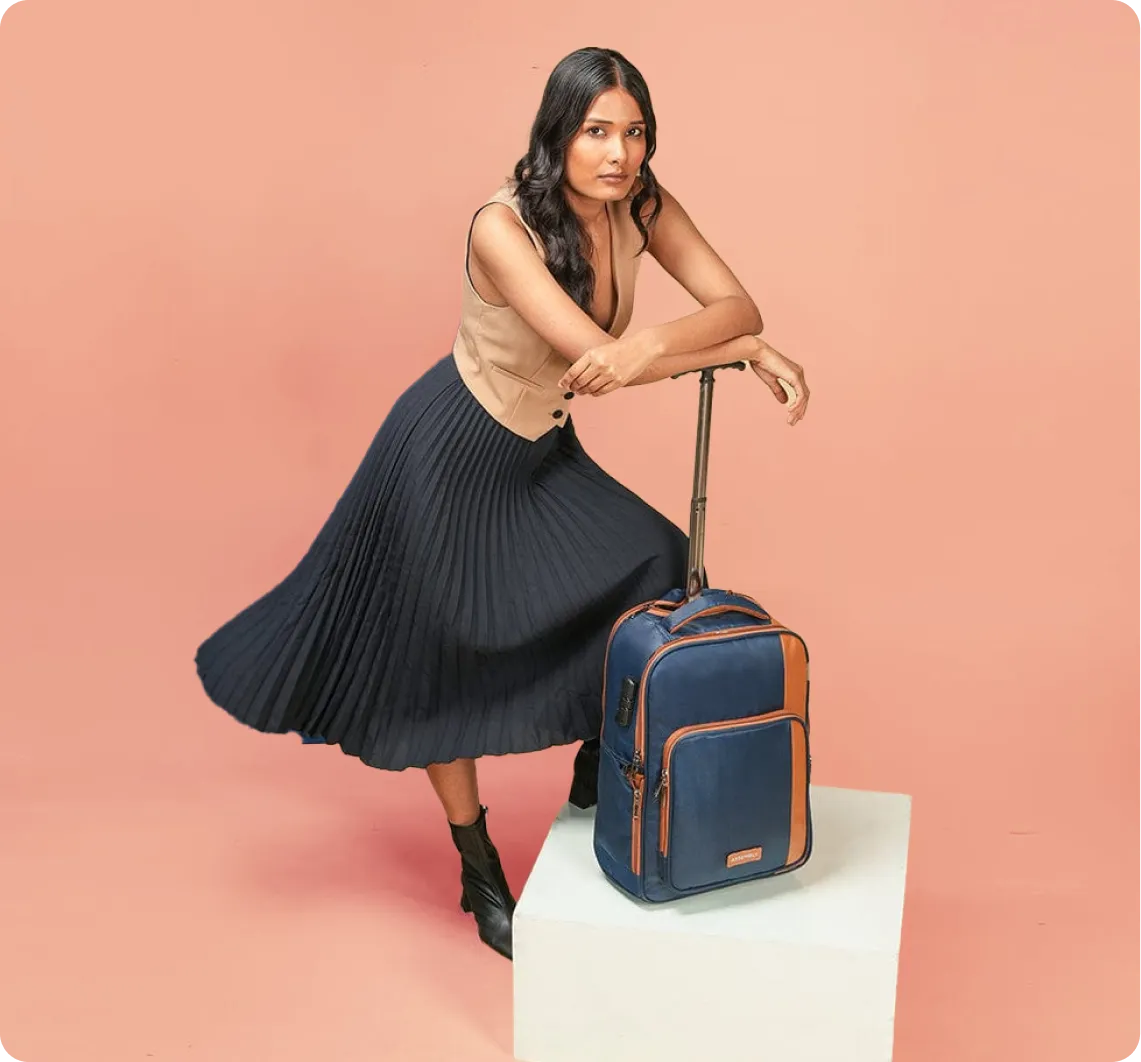 Woman with long dark hair wearing a sleeveless beige top, a dark pleated skirt, and black ankle boots, leaning on the handle of a blue and brown wheeled suitcase placed on a white pedestal against a peach background.