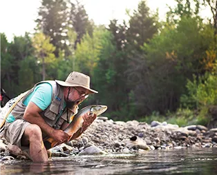 Man wearing hat holding a fish by a rocky riverbank surrounded by trees.