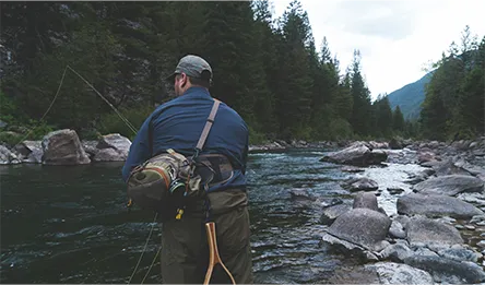 Man fly fishing in a rocky river surrounded by forested hills under cloudy sky.