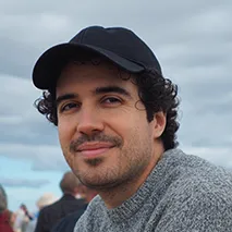 Smiling man with curly hair wearing a black cap and gray sweater outdoors under cloudy sky.