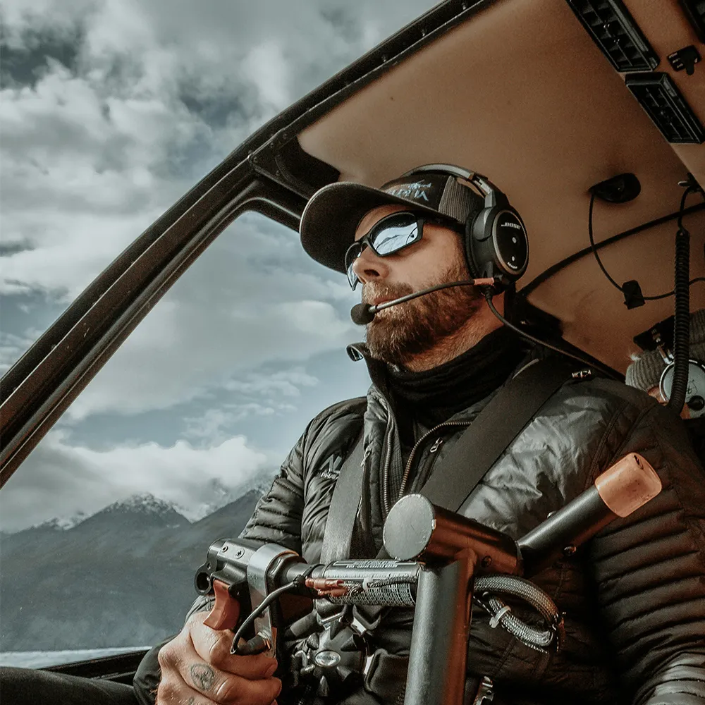 Bearded man wearing headset and sunglasses piloting a helicopter with mountain view outside.