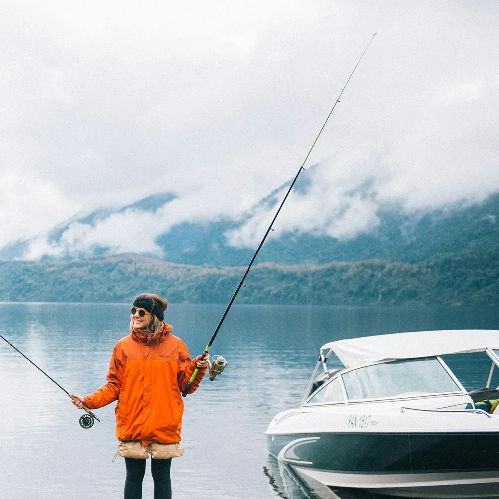 Person in orange jacket holding fishing rods near a boat on calm lake with misty mountains in background.