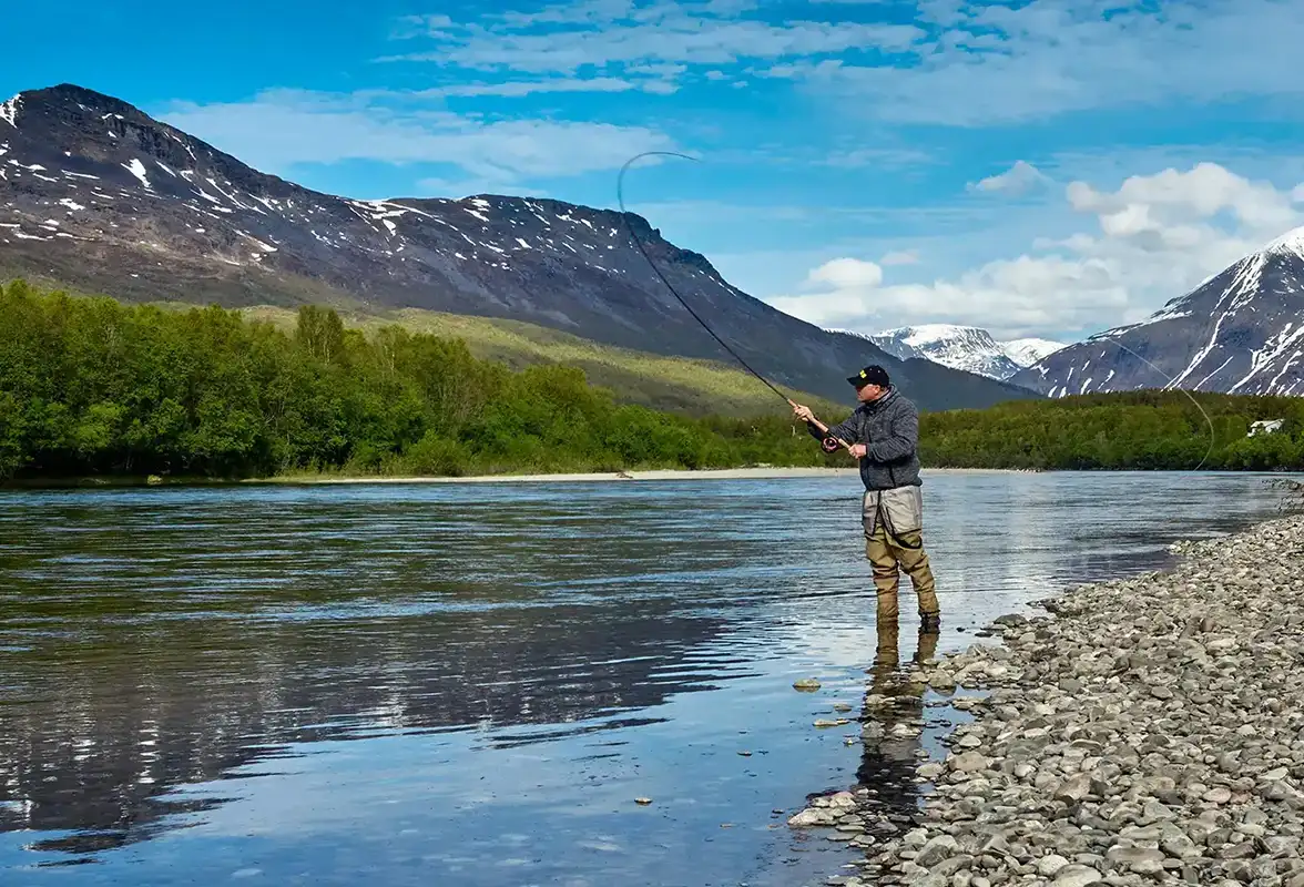 Fisherman fly fishing in a river surrounded by rocky shore, green forest, and snow-capped mountains.