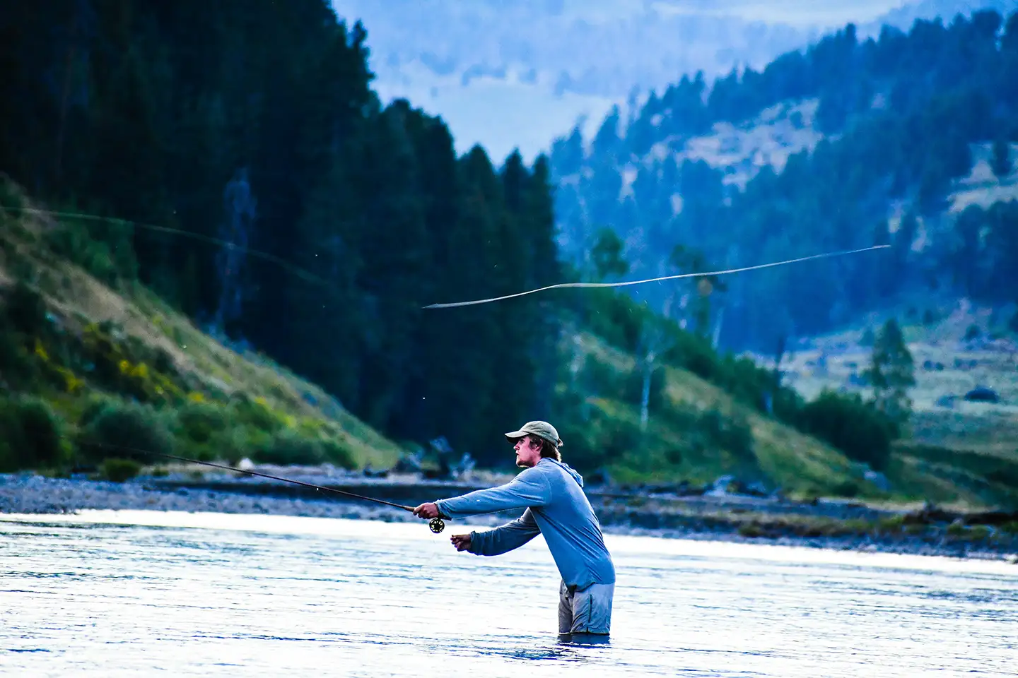 Man in a cap fly fishing waist-deep in a river with forested hills in the background.