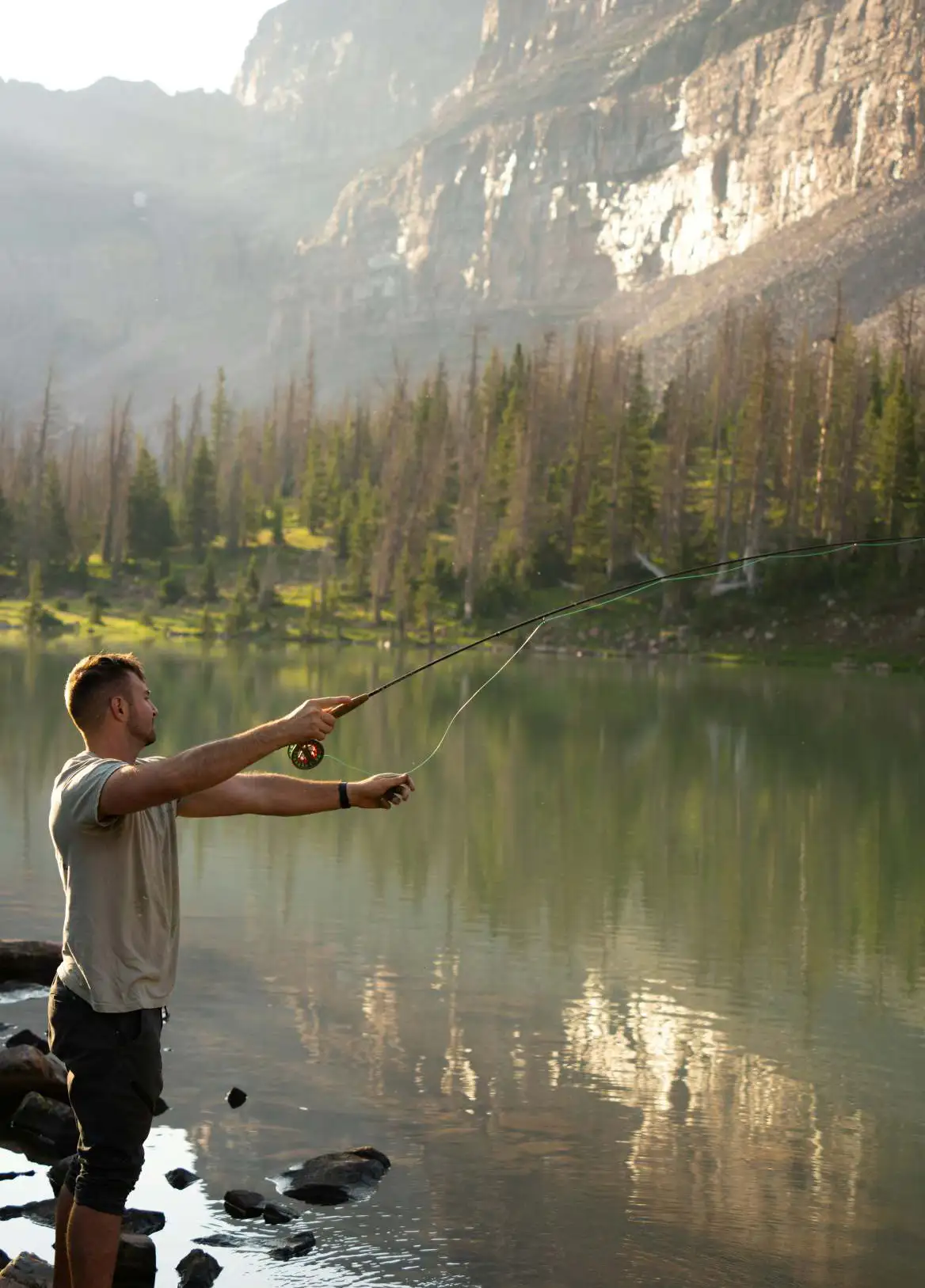 Man in green shoes fly fishing on a rocky shore of a mountain lake surrounded by trees.