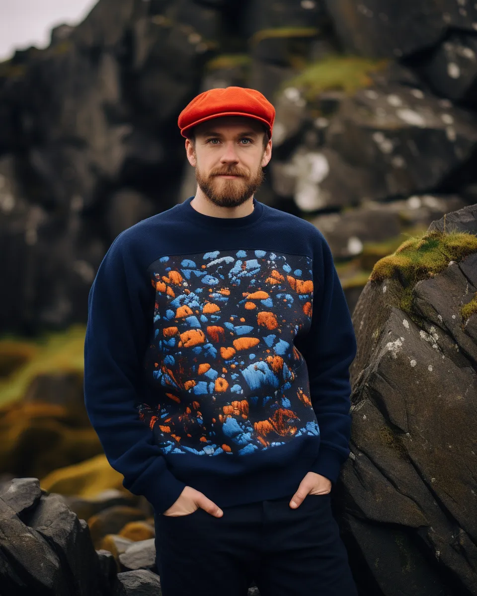 Man with beard wearing red cap and blue sweater with orange-blue rock pattern standing by mossy rocks.