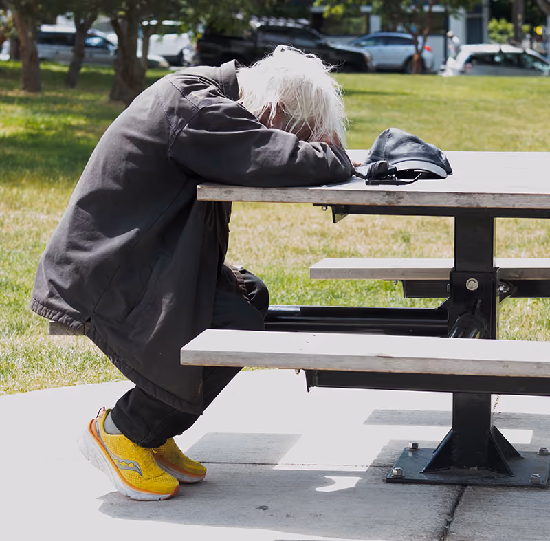 Elderly person with white hair resting head on arms on a park picnic table with yellow sneakers.