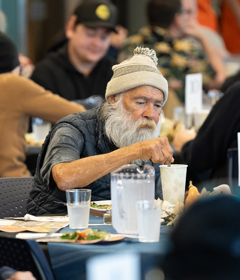 Elderly man with gray beard and knit hat eating at a crowded community meal table.