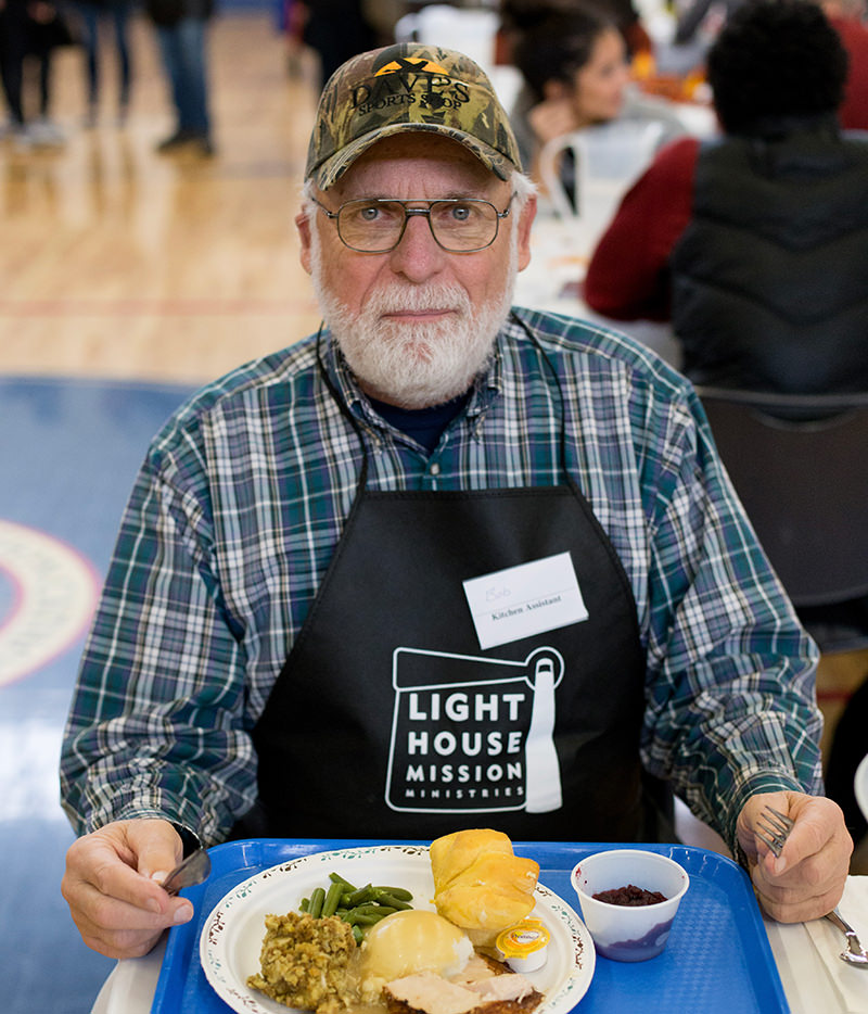 Elderly man in a camo cap and Light House Mission apron with a meal tray at a community dining event.