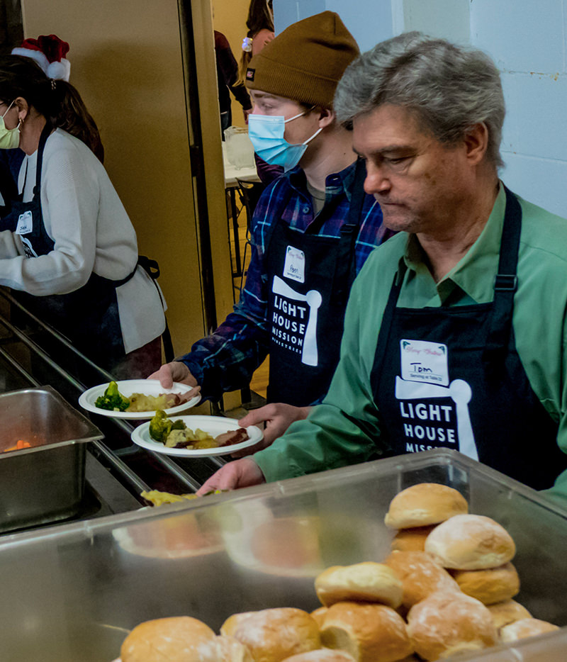 Volunteers wearing Light House Mission aprons serving food trays with rolls and vegetables.