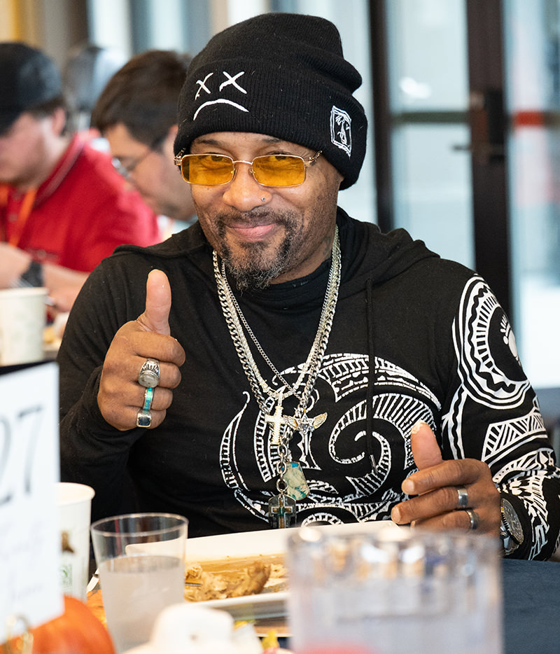 Man wearing a black beanie, yellow glasses, and patterned shirt giving a thumbs-up at a meal table.