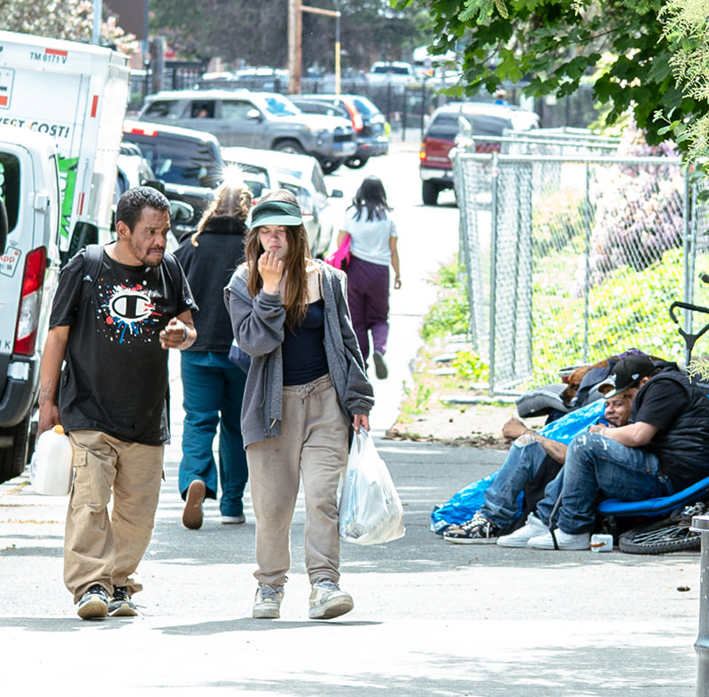 People walking on a city sidewalk past two men sitting by a fence surrounded by bags and belongings.