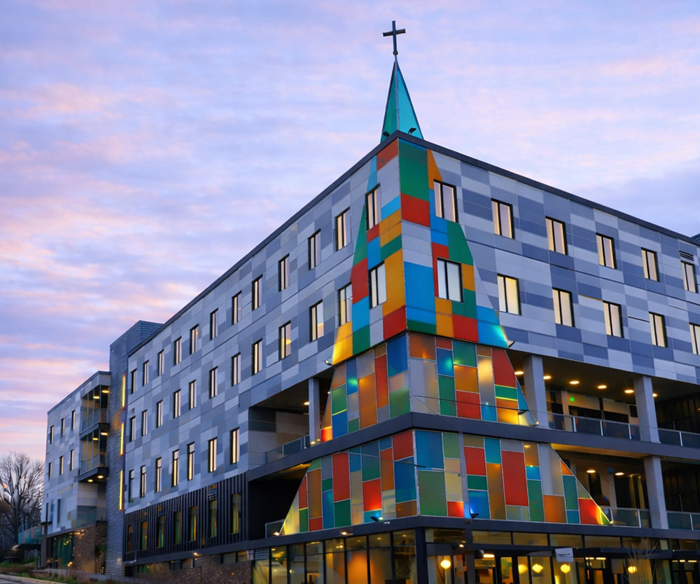 Modern building with colorful stained-glass cross-shaped tower topped by a black cross at sunset.