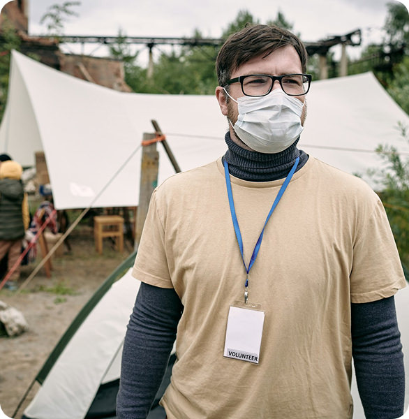 Masked volunteer wearing glasses and a beige shirt with a volunteer badge standing near a tent outdoors.