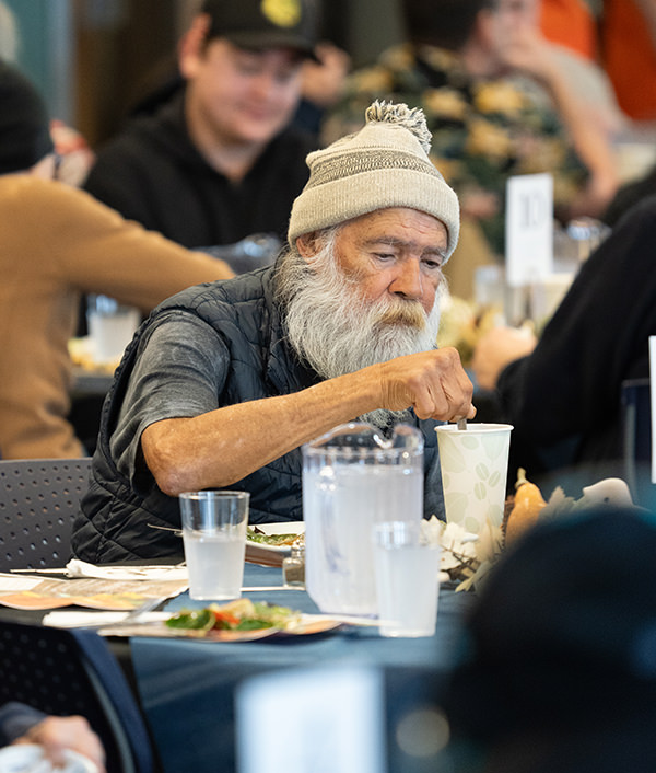 Elderly man with white beard and knit hat sitting at a table stirring a drink at a crowded dining area.