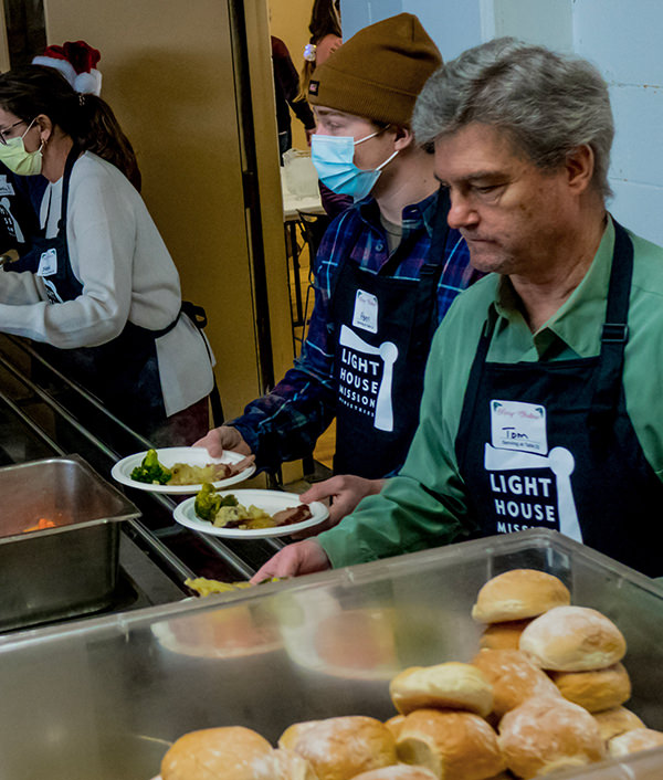 Volunteers wearing Light House Mission aprons serve food to people in line at a shelter or community meal.