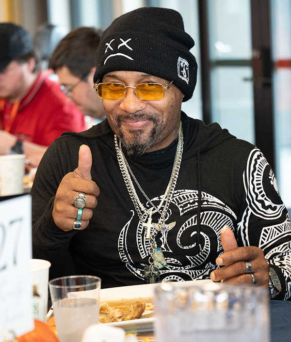 Man wearing a black beanie and yellow glasses giving a thumbs-up while sitting at a table with food.
