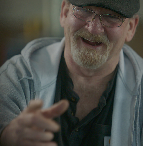 Smiling bearded man wearing glasses, a cap, a black shirt, and a gray hoodie reaching forward.