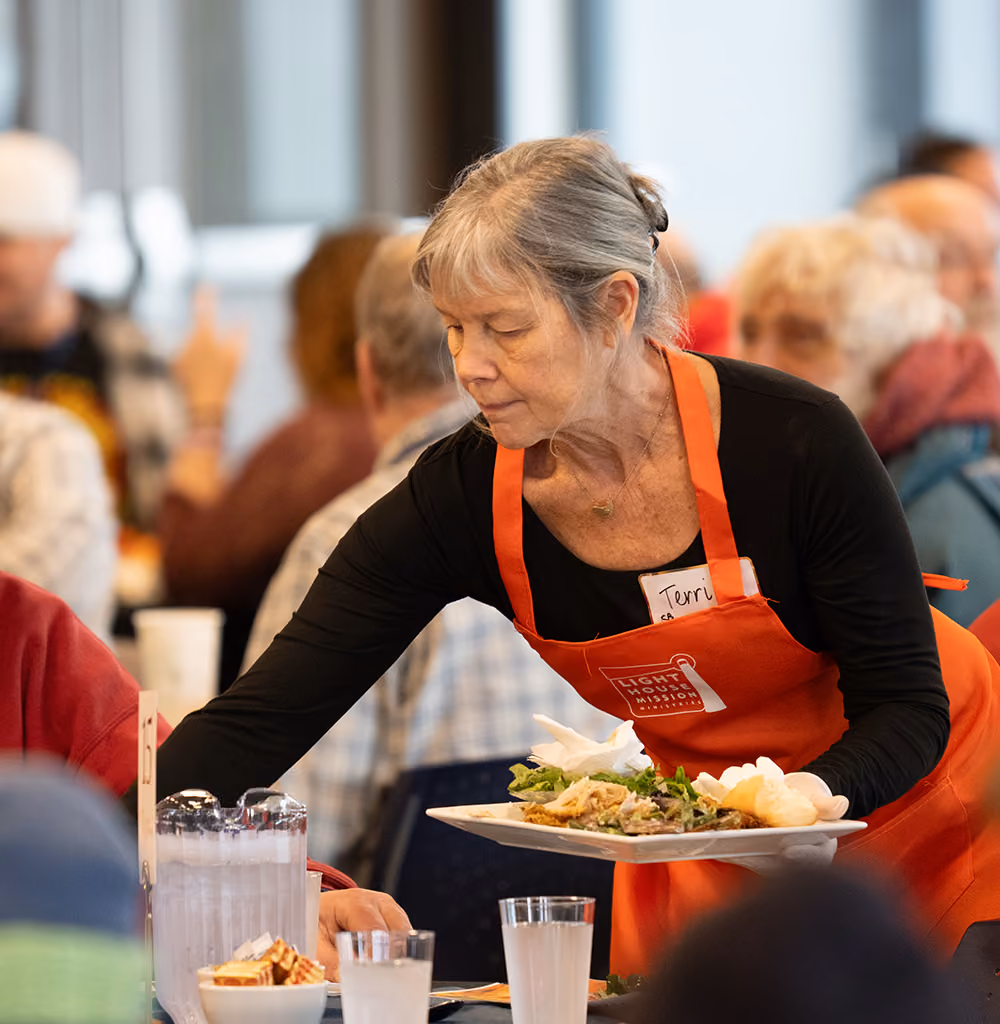Volunteer woman in an orange apron serving a meal on a plate to seated people at a dining table.