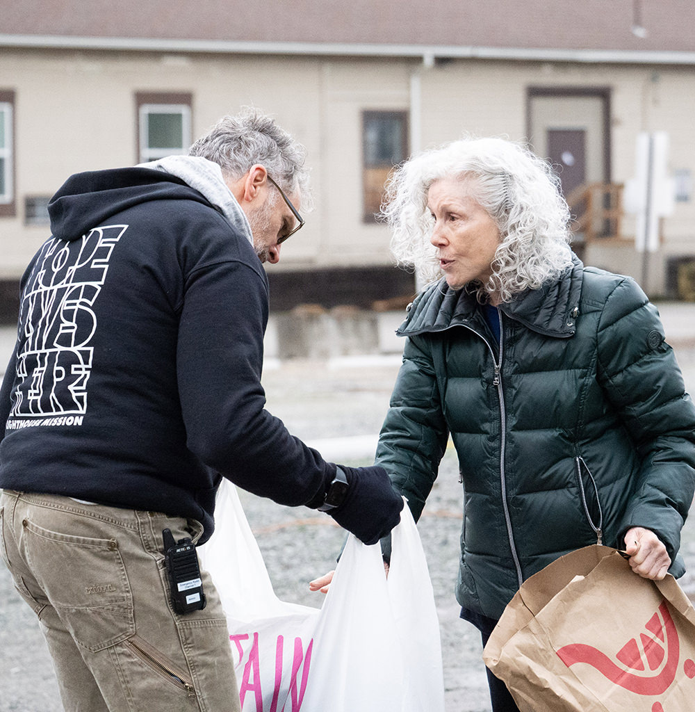 Man in black hoodie handing a white bag to a woman in green jacket holding a brown paper bag outdoors.
