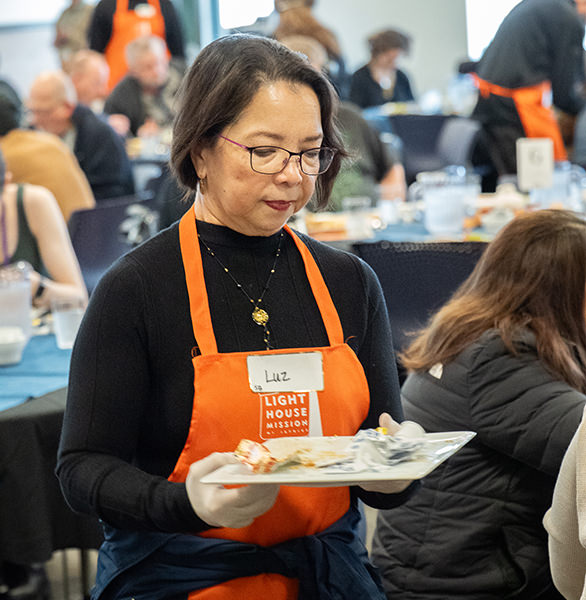 Woman volunteer in orange apron carrying a tray in a community dining area with people eating nearby.
