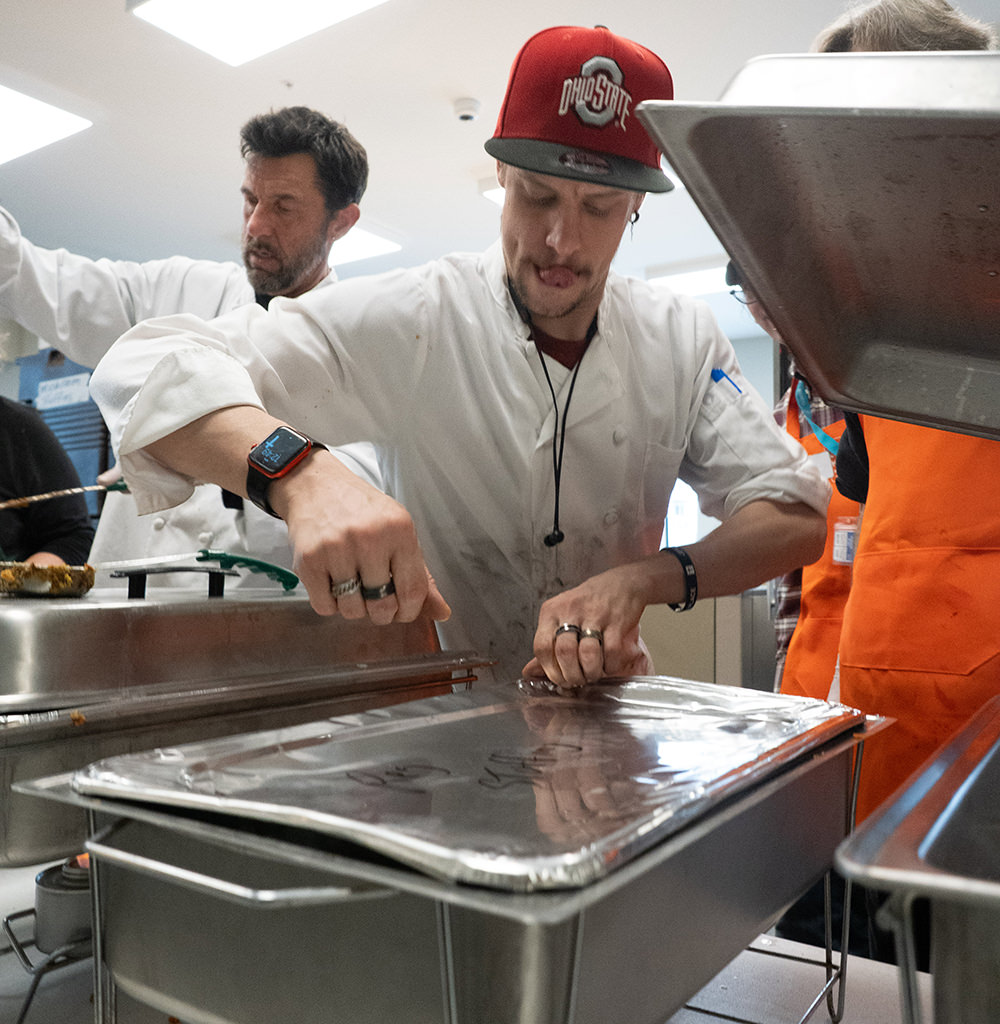 Man in chef coat and red Ohio State cap preparing food behind metal trays in a busy kitchen.