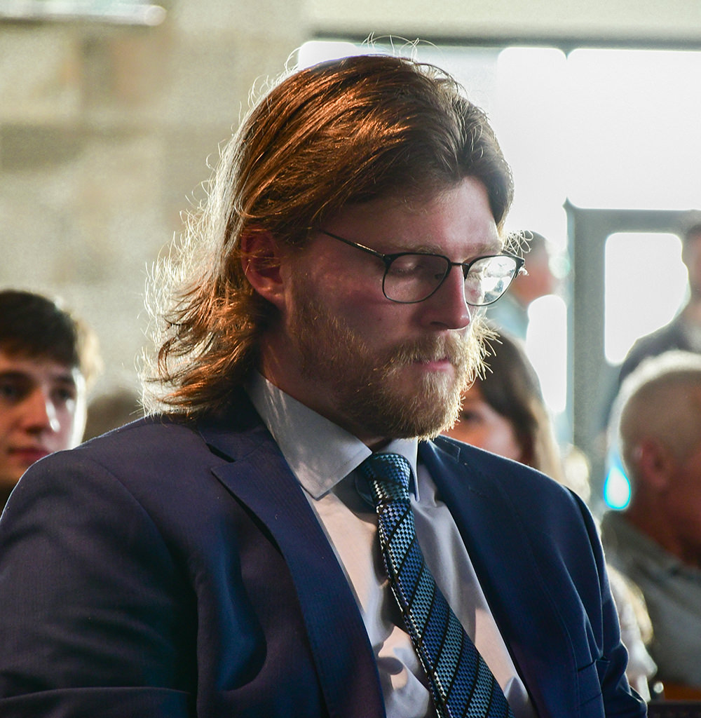 Man with glasses and beard in a blue suit and tie, eyes closed, sitting among people indoors.