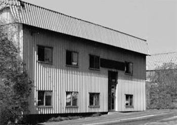 Black and white photo of a two-story corrugated metal building with six windows and a door.