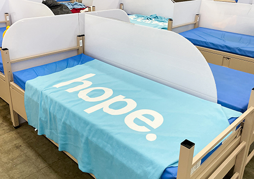 Bed in a dormitory with a light blue blanket labeled 'hope.' and blue sheets on a stretcher-style frame.