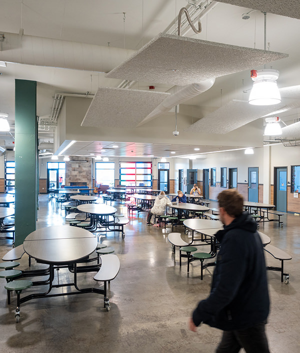 Spacious school cafeteria with round tables, a few people seated, and a person walking in foreground.