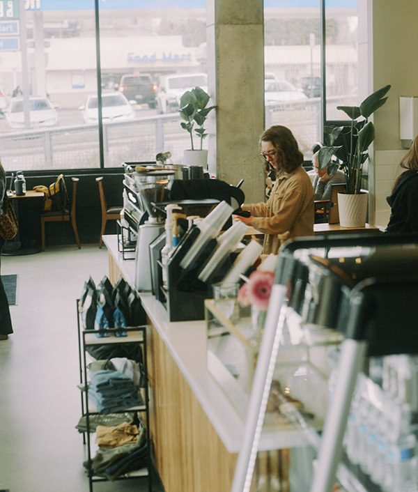 Barista preparing drinks behind counter in a bright coffee shop with plants and packaged goods.