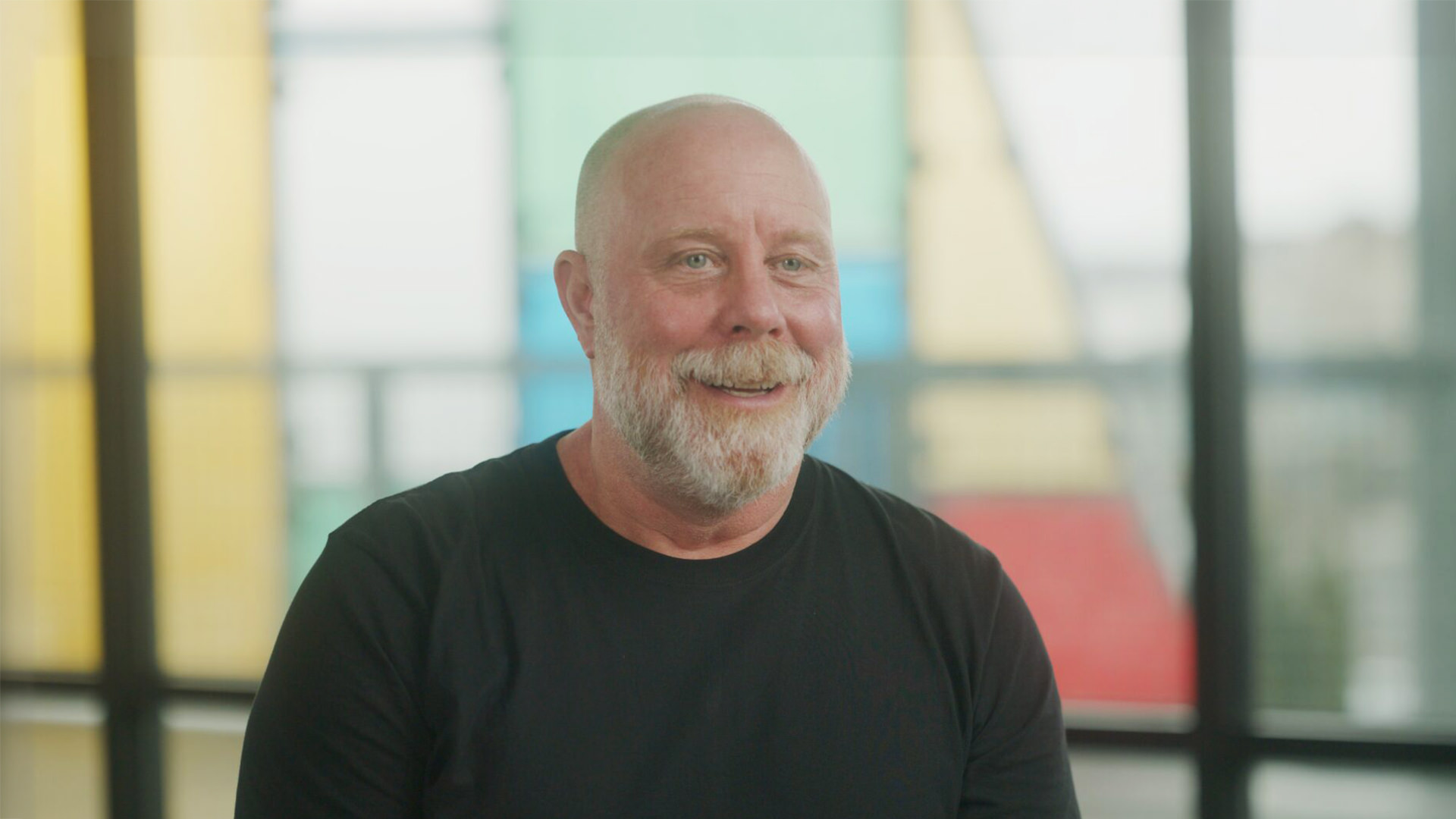 Smiling bald man with a white beard wearing a black shirt in front of colorful window panels.