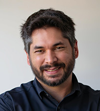 Reuben Evans with dark hair, beard, and mustache wearing a dark shirt against a plain background.